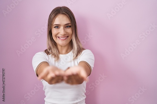 Blonde caucasian woman standing over pink background smiling with hands palms together receiving or giving gesture. hold and protection
