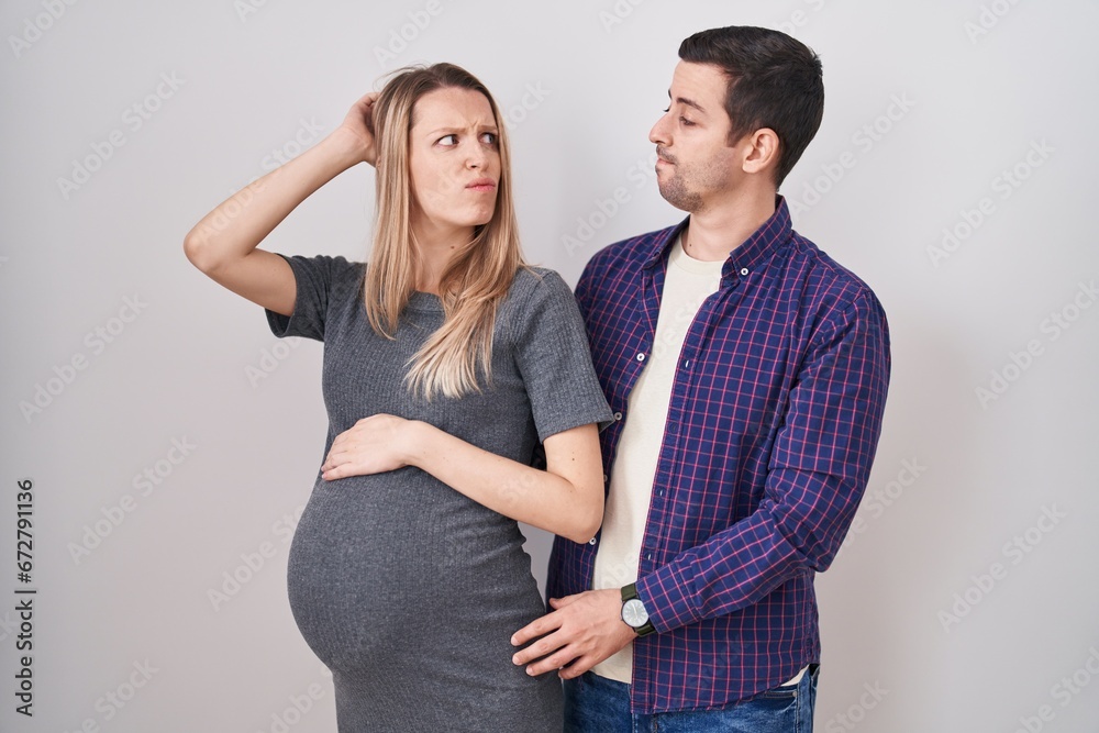 Young couple expecting a baby standing over white background confuse and wondering about question. uncertain with doubt, thinking with hand on head. pensive concept. Young couple expecting a baby standing over white background confuse and wondering about question. uncertain with doubt, thinking with hand on head. pensive concept.