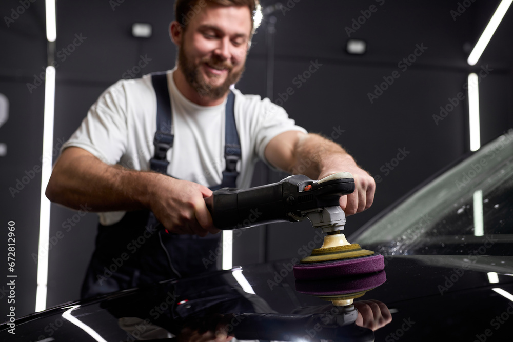 Auto Mechanic Use polishing tool for Buffing and polishing black car In auto repair shop, side view. Confident male worker in uniform at work place concentrated. focus on polisher
