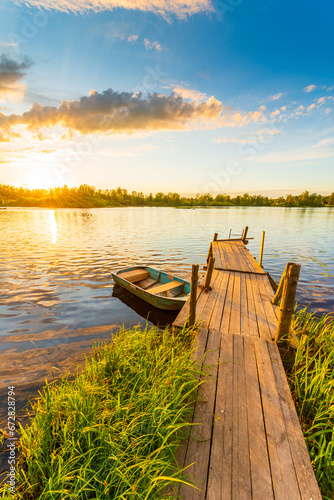 Sunset over the lake. Wooden village bridge with a boat. Cloudy sky. Ripples in the water. Reed thickets. View from the bridge. Russia, Europe. Vertical shot.