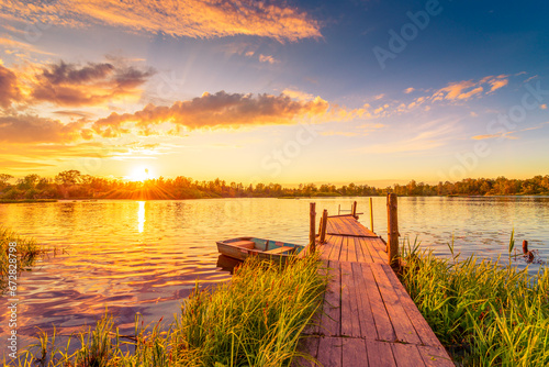 Fototapeta Naklejka Na Ścianę i Meble -  Sunset over the lake. Wooden village bridge with a boat. Cloudy sky. Ripples in the water. Reed thickets. View from the bridge. Russia, Europe.