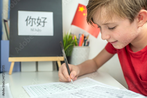 An 11 year old child is learning Chinese. a happy European boy sits at a desk, in front of him is a Chinese flag, on the desk is the hieroglyph “Hello” in Chinese, the boy is doing his homework  photo