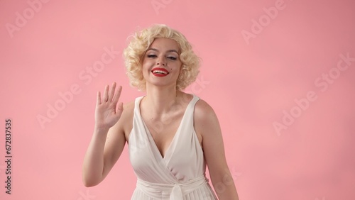 A woman dressed as waves her hand in greeting. A woman in a white dress and white wig with red lipstick on her lips in the studio on a pink background.