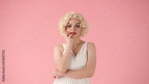 Portrait of a beautiful woman smiling sensually, touching her lips coquettishly with her fingers. Woman in the image of , in white wig, dress in studio on pink background.