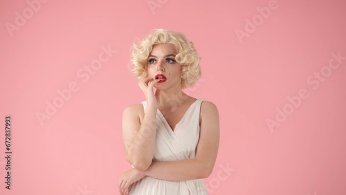 Woman in the image of , dreamily thinking about something, coquettishly putting her fingers to her lips. Woman with bright makeup in white wig and white dress isolated on pink background