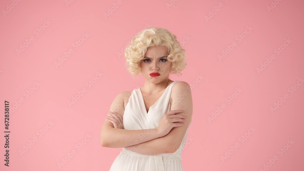 A young woman stands offended, pouting her lips with red lipstick and crossing her arms over her chest. Portrait of a woman in the image of in a studio on a pink background. A young woman stands offended, pouting her lips with red lipstick and crossing her arms over her chest. Portrait of a woman in the image of in a studio on a pink background.