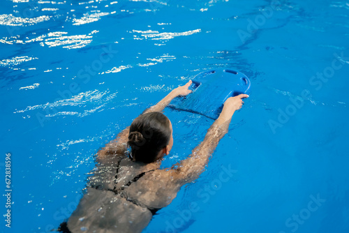 happy girl learning how to swim in the swimming pool, holding blue pool board in the water