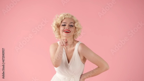 A young woman in the image of points her index finger at you. Woman with bright makeup and in white dress in studio on pink background close up.