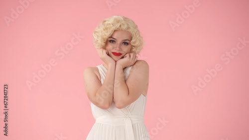Portrait of young cheerful woman in wig, white dress and with red lipstick on lips in studio on pink background. Woman in the image of , holding her face in the palms of her hands.