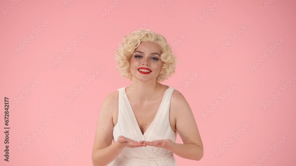 A young woman with bright makeup holds empty palms in front of her. Woman in the image of merilyn monroe shows empty space in studio on pink background. A young woman with bright makeup holds empty palms in front of her. Woman in the image of merilyn monroe shows empty space in studio on pink background.