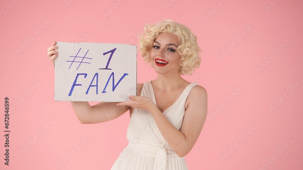 A joyful woman holds up a placard that reads fan number one. Woman looking like in studio on pink background close up. A joyful woman holds up a placard that reads fan number one. Woman looking like in studio on pink background close up.