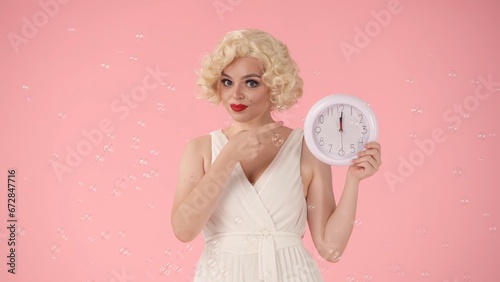 Woman holding a white round wall clock in her hand and pointing her index finger at the time. Woman looking like in studio surrounded by soap bubbles on pink background. Time concept.