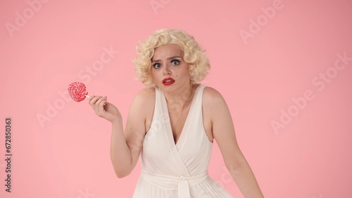 Woman holding a lollipop on a stick and shrugging her shoulders. Woman looking like in studio on pink background.
