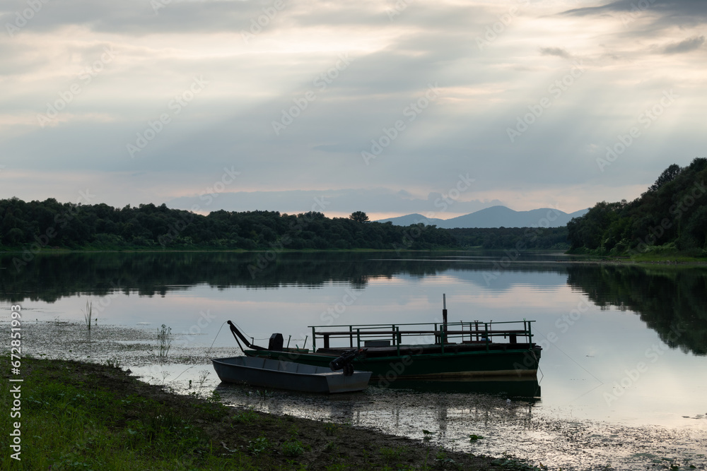Fototapeta premium Landscape with moored boats in river against mountain silhouette in distance and sun rays coming from clouds