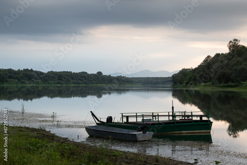 Fototapeta Naklejka Na Ścianę i Meble -  Landscape with moored boats in river against mountain silhouette in distance and cloudy sky