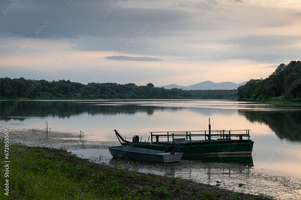 Landscape with moored boats in river against mountain silhouette in distance and cloudy sky
