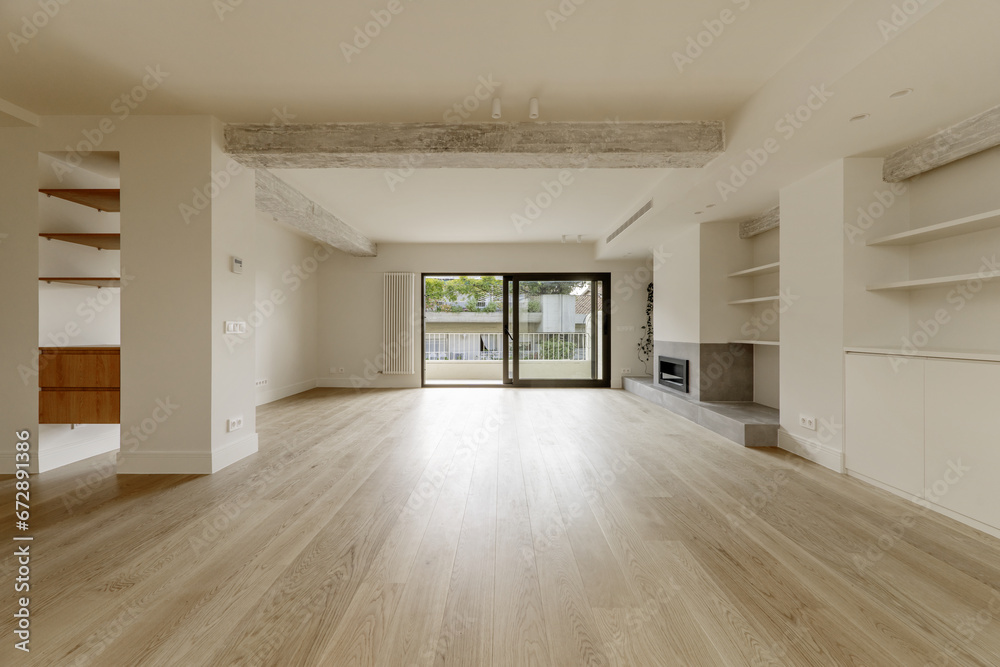 Large empty living room with custom wooden shelving on the walls, exposed concrete beams, white oak wooden floors, cream painted walls and access to a terrace with glass doors