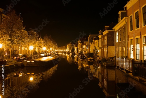 Fleet of boats in a canal in front of a picturesque row of houses at night: Leiden © Wirestock