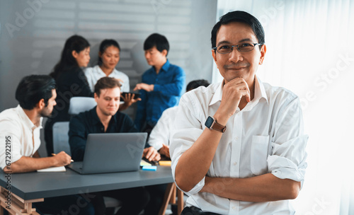 Confidence and happy smiling businessman portrait with background of his colleague and business team working in office. Office worker teamwork and positive workplace concept. Prudent