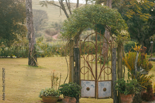 Vintage garden gate entwined with climbing plants leads to a tranquil meadow, flanked by potted shrubs and rustic charm.