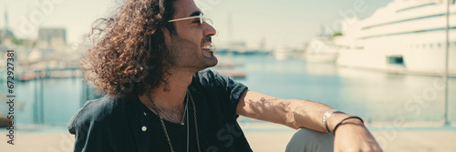 Young italian guy with long curly hair and stubble sits in harbor and looks at ships and yachts photo