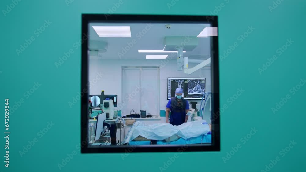 A look through the window in the surgery room door. Male doctor wearing x-ray protective suit is inside the room setting equipment.