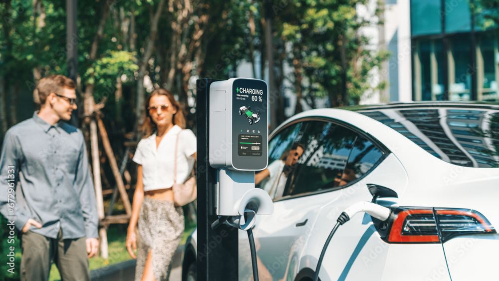 Young couple travel with EV electric car charging in green sustainable city outdoor garden in summer shows urban sustainability lifestyle by green clean rechargeable energy of electric vehicle innards - obrazy, fototapety, plakaty 