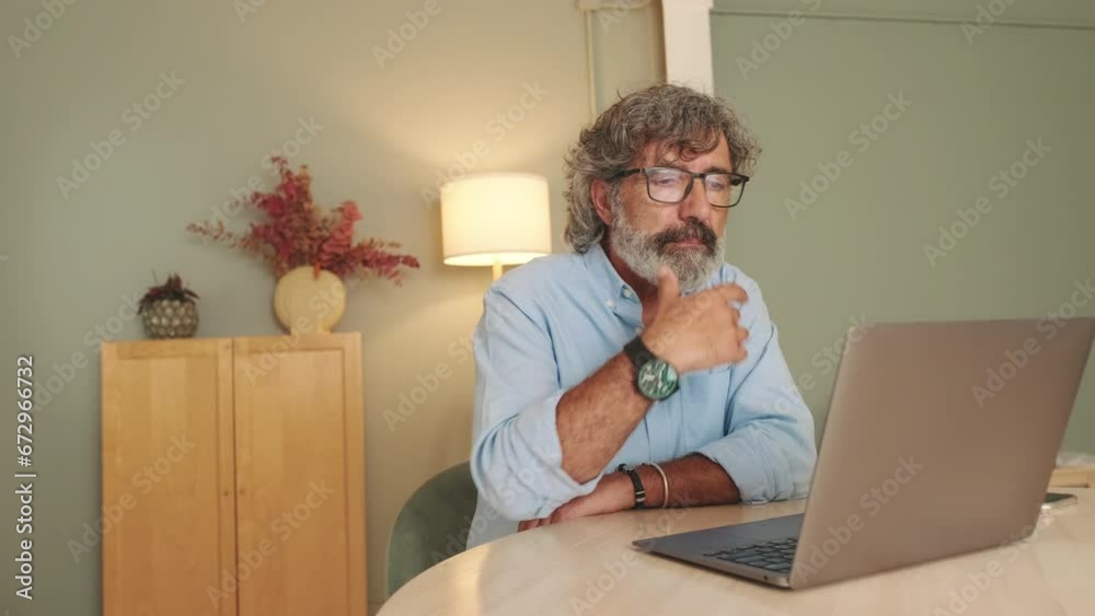 Pensive middle-aged man in glasses sitting in the living room working on laptop Pensive middle-aged man in glasses sitting in the living room working on laptop