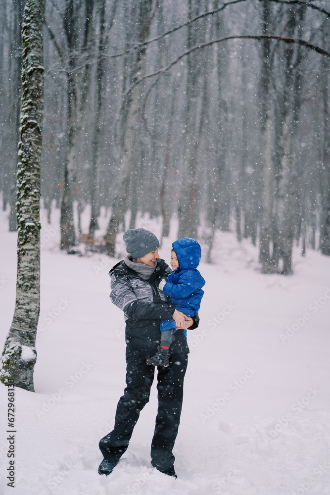 Smiling mother with a little girl in her arms stands under snowfall in the forest