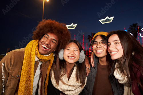 Four joyful friends posing smiling selfie mobile looking at camera standing hugging at christmas winter night outdoor. Multi-ethnic group of cheerful people in amusement park having fun together.