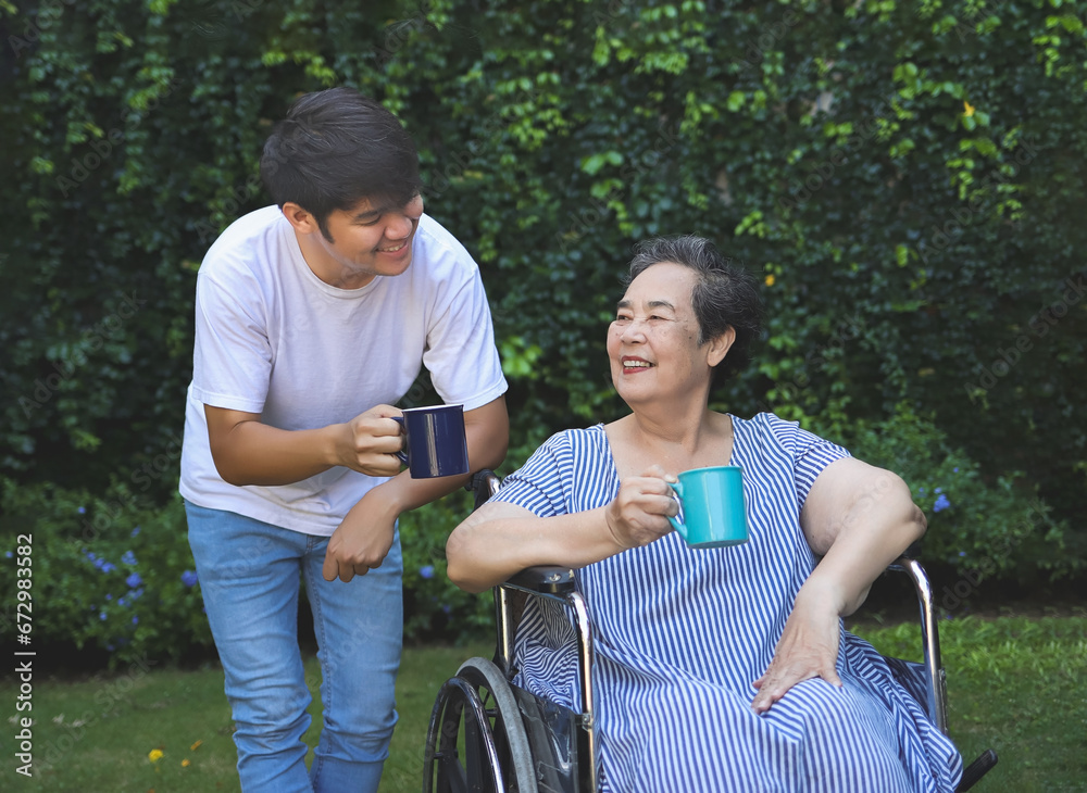 Obraz premium Asian senior woman sitting on wheelchair, drinking coffee or tea with her son in the garden. smiling happily.