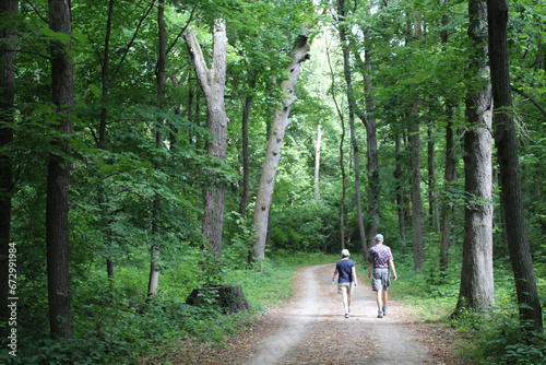 Older couple walking on the Des Plaines River Trail among tall trees at Camp Ground Road Woods in Des Plaines, Illinois