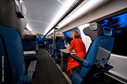 Young female traveler sitting in the modern train wagon near window during evening time using smartphone, back view. Concept of urban journey, transportation, travel and trip. Copy space for text.