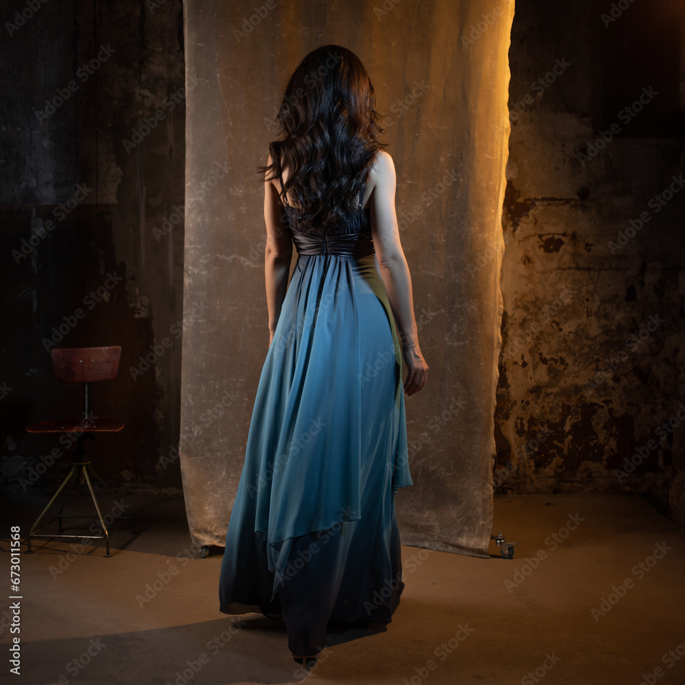 An elegant and beautiful brunette in a chic evening dress, a blue dress with a long flowing skirt to the floor. photo in dark colors on a textured wall, warm light