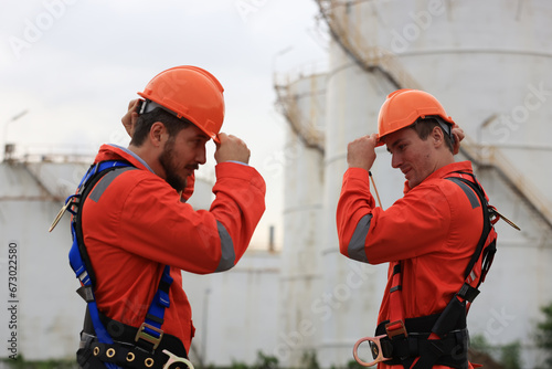 Two maintenance engineers in safety uniforms with orange hard hats and safety harnesses are ready to work at Oil tank storage.