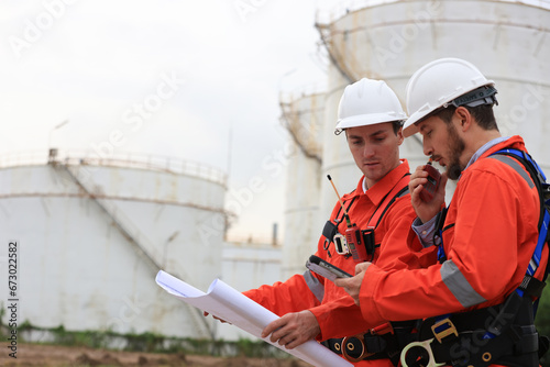 Team Engineers wearing safety harnesses working and discussing with blueprint at the Oil Refinery storage tanks. photo