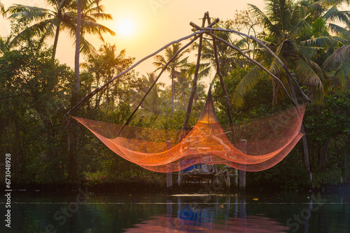 A traditional Chinese fishing net is raised by a crane structure in the sunset on a lake, Backwaters, Kerala state, India