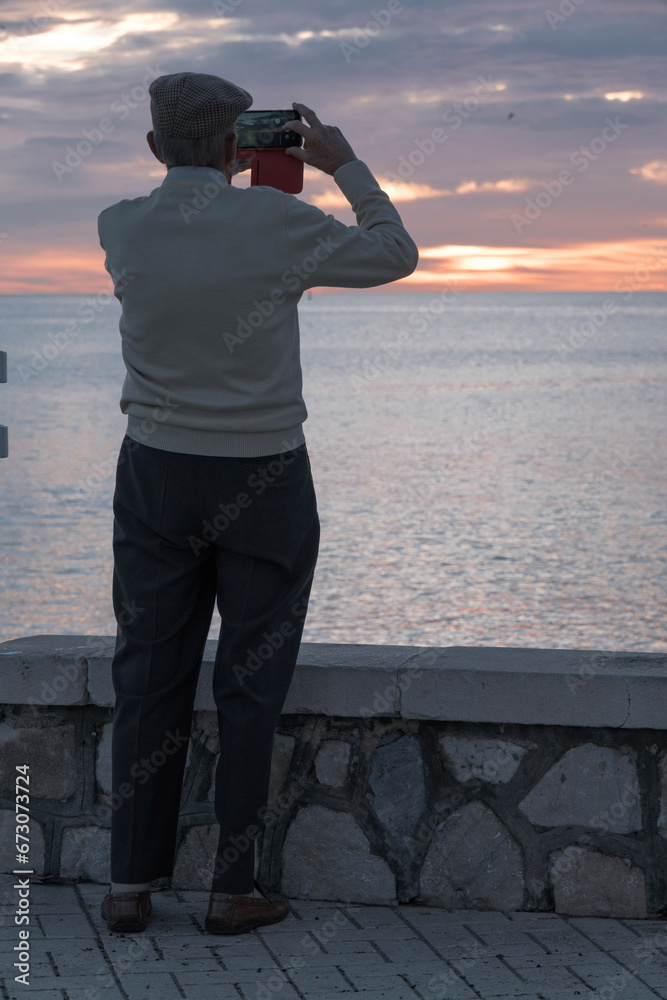 Obraz premium Hombre mayor de espaldas, paseo marítmo de Máaga, anciano haciendo fotos a la playa al amanecer, primeros rayos de sol