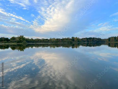 White cumulus clouds in a blue sky are beautifully mirrored in a blue lake against the background of an autumn forest and a suburban cottage village on a beautiful warm day. High quality FullHD