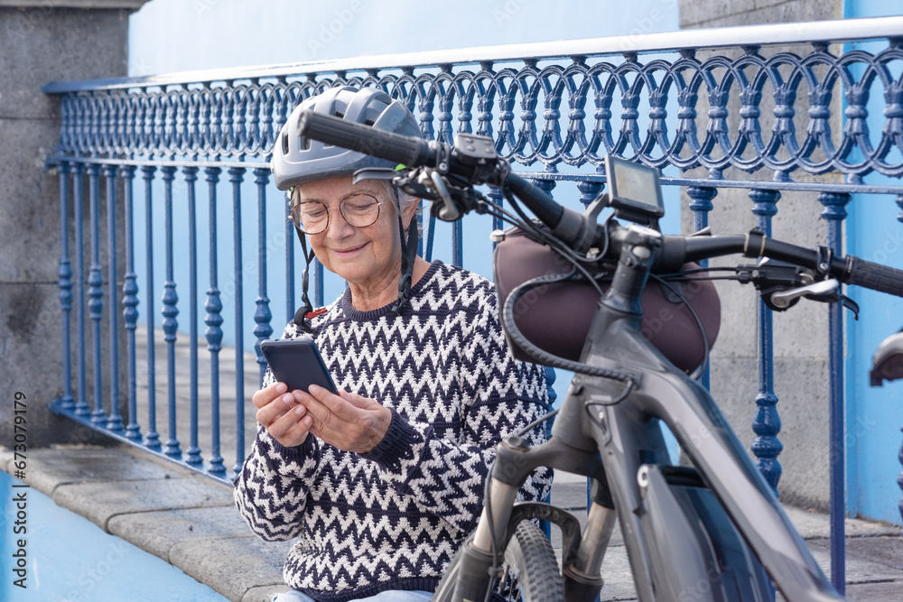 Smiling senior cyclist woman wearing bike helmet sitting close to her bicycle using mobile phone typing a message. Relaxed pensioner female enjoying free time and healthy lifestyle