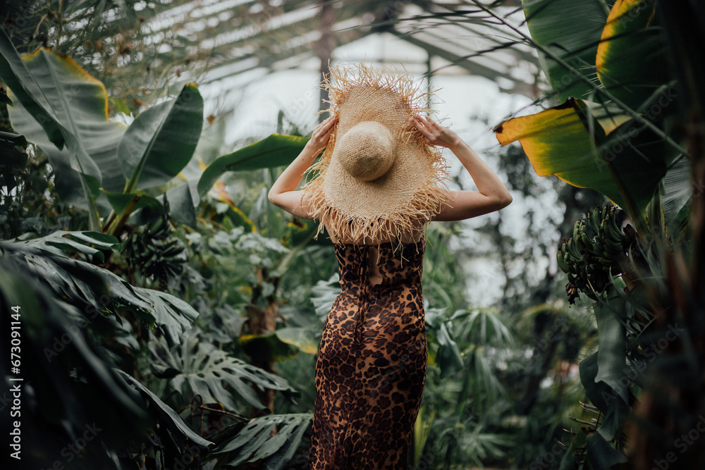 Obraz premium Back view of woman tourist in straw hat and leopard dress standing in palm greenhouse 