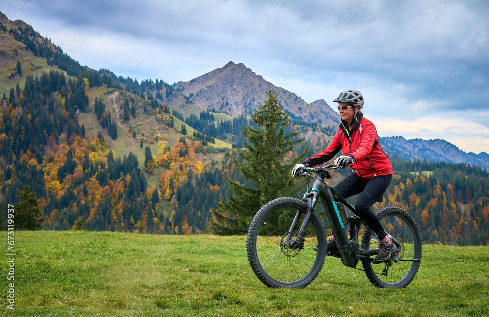 pretty senior woman riding her electric mountain bike in autumn and enjoying the spectacular view over the Allgau and Bregenz Forest alps near Steibis, Bavaria, Germany