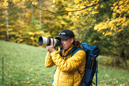 Hiker with backpack is wearing yellow jacket and photographing at his camera. Nature or landscape photographer in autumn forest