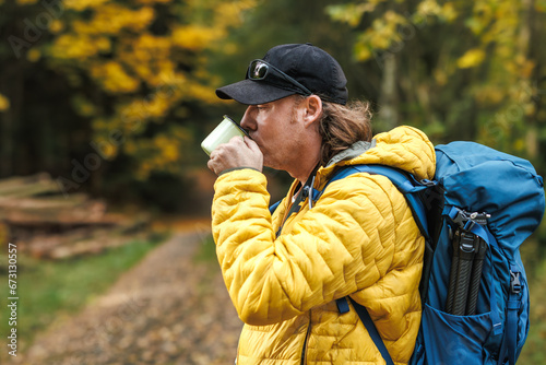 Hiker with backpack and yellow jacket drinking hot tea from travel mug in autumn forest. Refreshment and resting during outdoor hiking