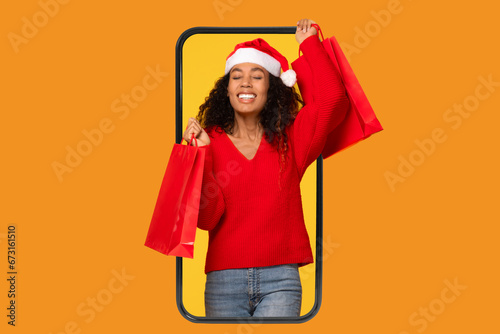 Excited black lady wearing Santa hat holding shopper bags in phone screen, orange backdrop