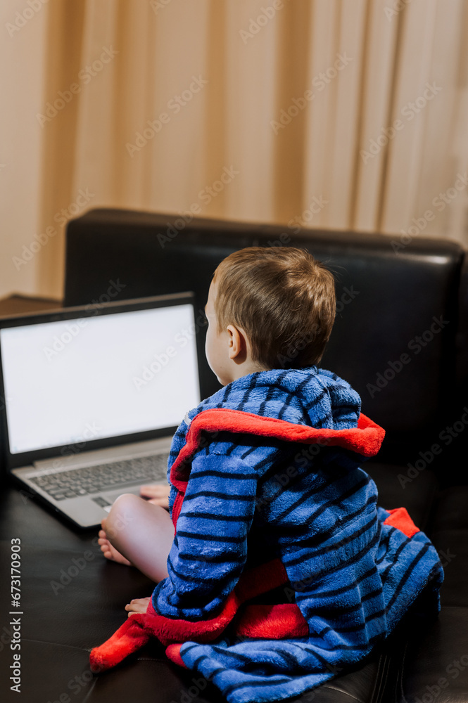 A little boy, a child in pajamas sits on a black leather sofa and looks at a laptop, playing games, watching interesting movies via the Internet. Modern childhood concept, copy space.
