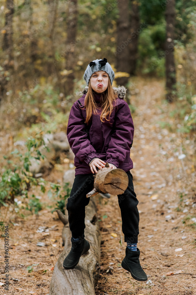 Beautiful smiling happy girl, child makes faces, grimaces, shows tongue, sitting on a log in the forest in autumn. Photography, portrait.