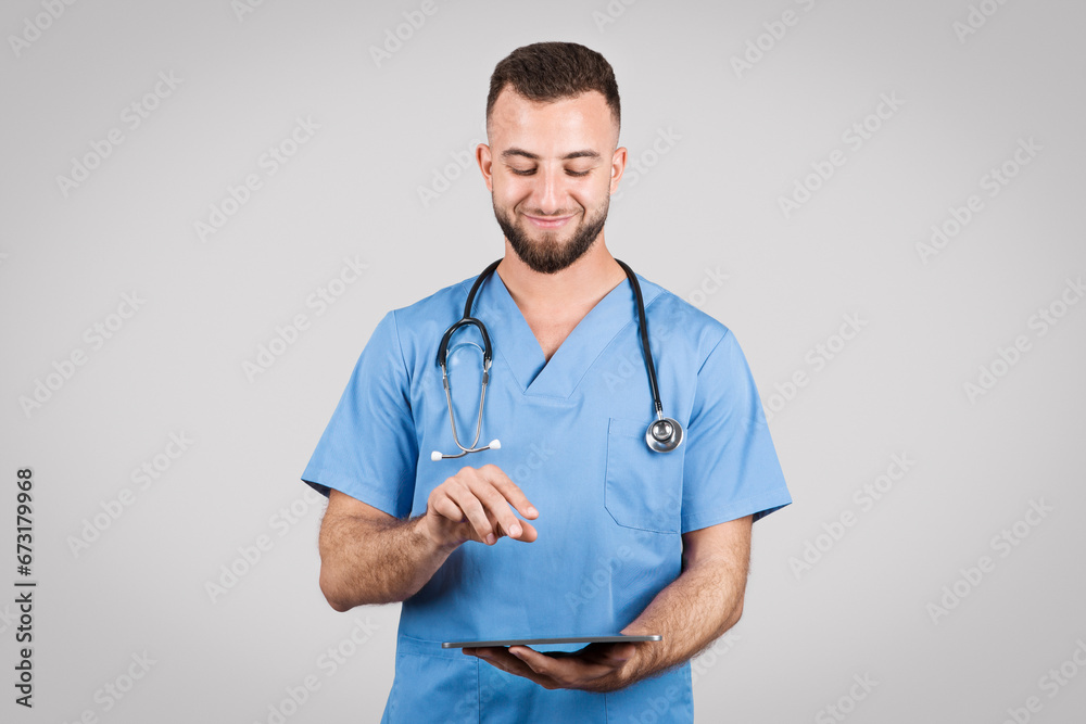 Doctor man in blue uniform using tablet and smiling on grey background Doctor man in blue uniform using tablet and smiling on grey background