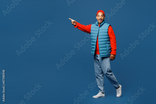 Full body side view surprised young man of African American ethnicity wear padded vest red hat walk go point aside isolated on plain dark royal navy blue background studio portrait. Lifestyle concept.