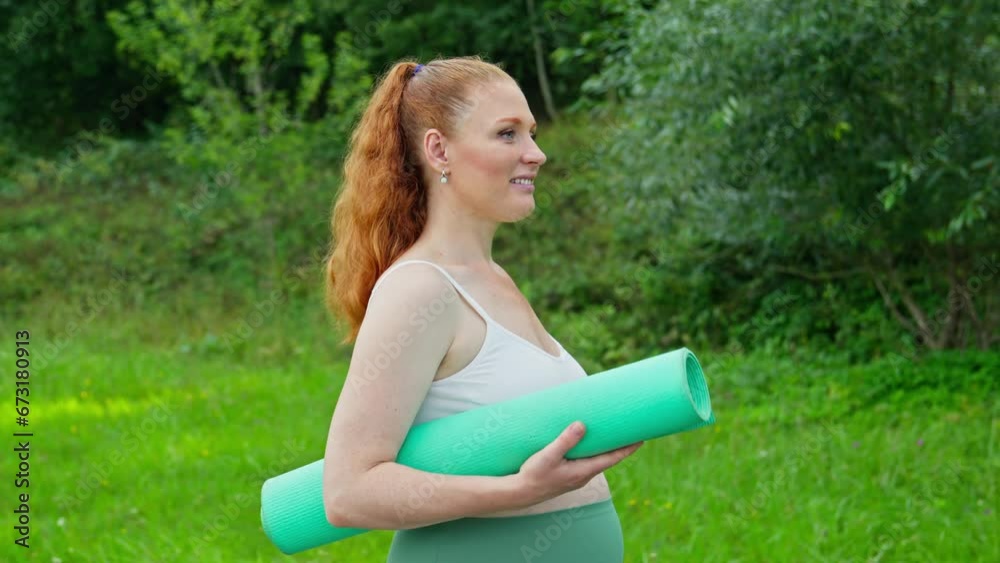 Redhead pregnant woman walking in park with yoga mat. Expecting female in park. Maternity prenatal care and pregnancy concept. Tracking shot. Healthy lifestyle. 4K, UHD Redhead pregnant woman walking in park with yoga mat. Expecting female in park. Maternity prenatal care and pregnancy concept. Tracking shot. Healthy lifestyle. 4K, UHD